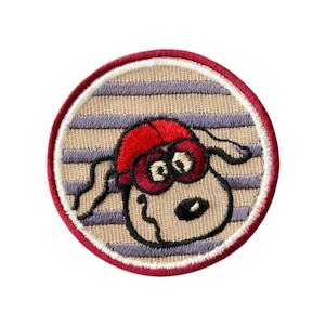 Op de afbeelding: Gestikt patch met een cartoon hond die een rode hoed met witte strepen draagt. De hond heeft bruine ogen en een zwarte neus. Het patch is rond en heeft een beige achtergrond met paarse strepen.