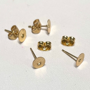 Könnte beinhalten: Eine Sammlung goldfarbener Ohrstecker und -verschlüsse. Die Ohrstecker haben eine flache, runde Scheibe vorne und einen dünnen Stift. Die Verschlüsse sind schmetterlingsförmig. Diese Komponenten dienen der Schmuckherstellung.