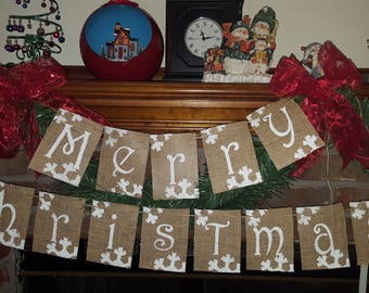 May include: A burlap banner with the words "Merry Christmas" in white lettering. The banner is decorated with white snowflakes and is hanging on a fireplace mantel. There are red bows and greenery on either side of the banner.