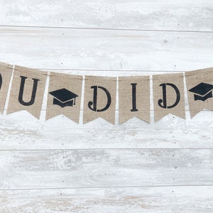 May include: A burlap banner with the text "YOU DID IT" in black lettering. The banner has graduation caps between the words.