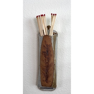 May include: A wall-mounted ceramic match holder with a speckled light gray finish and a central brown panel featuring a dark brown botanical leaf imprint. The holder contains several wooden matches with red tips, arranged vertically in the opening.