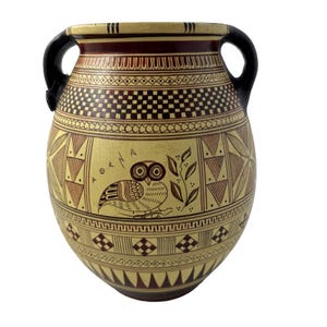 May include: A tan and brown ceramic vase with black handles. The vase features geometric patterns and an owl illustration. The word "AOKENA" is written on the vase. The vase is a decorative item.