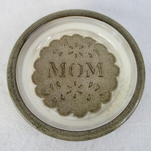 Op de afbeelding: Een keramische plaat met een grijze rand en een wit interieur. Het midden heeft een grijs, geschulpte ontwerp met het woord "MOM". Het ontwerp bevat bloem- en druppelvormen.