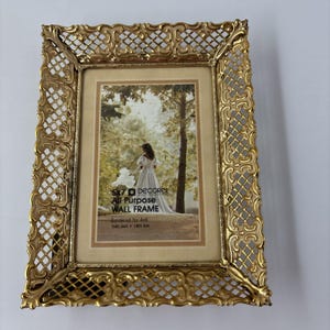 Puede incluir: Un marco de fotos rectangular de metal dorado con un borde de celosía ornamentado. El marco contiene una tarjeta con el texto 5x7 DECOREL All Purpose WALL FRAME Bordered for 4x6 1505 Gold 1805 Oak y una imagen de una persona con vestido blanco.