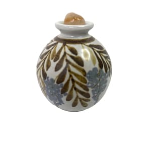 May include: A small, round ceramic jar with a white base, adorned with brown leaf and blue floral patterns. The jar has a narrow neck and is topped with a light brown stone. The jar is likely used for decorative purposes.
