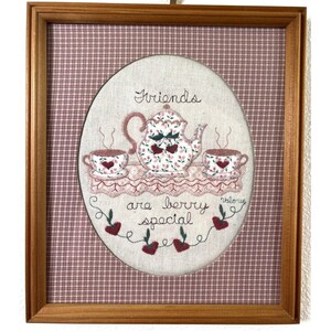 Può includere: Un ricamo incorniciato raffigurante una teiera e due tazze da tè su un centrino di pizzo, decorato con ciliegie rosse e foglie verdi. Il testo recita Friends are berry special. Lo sfondo è un tessuto a quadri rossi e bianchi in cornice di legno.