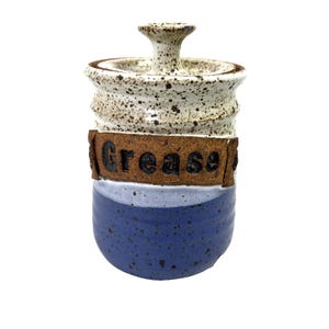 Puede incluir: Un tarro de grasa de cerámica hecho a mano con tapa. El tarro es cilíndrico con una base azul, una sección media moteada blanca y gris, y una banda marrón con la palabra "Grease" en letras negras. La tapa tiene un pomo.