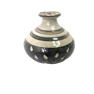 May include: A small, handcrafted ceramic vase with a rounded body and a narrow neck. The vase features alternating bands of cream and dark gray, with white, teardrop-shaped accents. The vase is a decorative item.