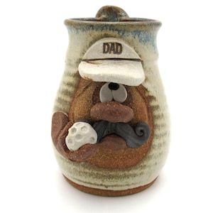 Puede incluir: Taza de cerámica con temática de golf. La taza es de color marrón y beige con la cara esculpida de un golfista que lleva un sombrero con la inscripción "DAD". El golfista sostiene una pelota de golf y tiene un bigote gris oscuro.