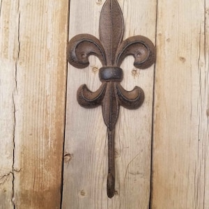 May include: A cast iron hook in the shape of a fleur-de-lis, painted in a dark brown color, mounted on a wooden wall.