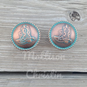 May include: Two copper-toned, round cabinet knobs with a turquoise rope-like border. Each knob features a detailed image of a cowboy boot. The knobs are set against a weathered wood background.
