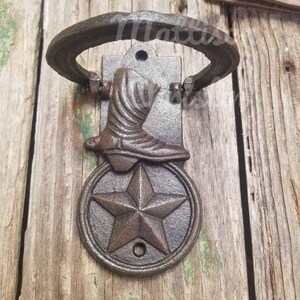 Op de afbeelding: Een rustieke gietijzeren deurklopper met een cowboylaarsontwerp boven een vijfpuntige ster in een cirkelvormig frame. De klopper heeft een gebogen handvat en is gemonteerd op een rechthoekige achterplaat, allemaal in een verweerde bronzen afwerking.