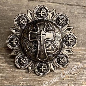 May include: Silver-toned metal concho with a cross design in the center. The concho is surrounded by a decorative border with smaller crosses.