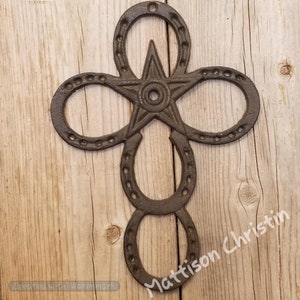 May include: A rustic metal cross made from four horseshoes with a star in the center. The cross is painted in a dark brown color and has a distressed finish.