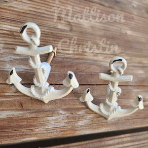 May include: Two white metal anchor-shaped drawer pulls with black accents. The anchors have rope details and are mounted on a wooden surface.