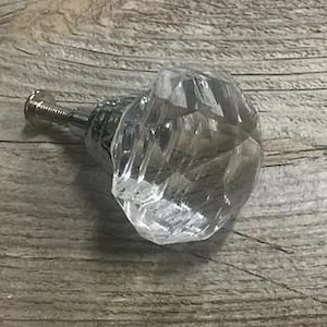 May include: A clear, faceted glass cabinet knob with a chrome-colored metal base and screw. The knob is diamond-shaped and reflects light, lying on a weathered wooden surface.