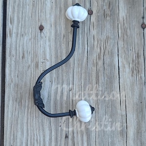 May include: Black metal hook with two white ceramic finials. The hook is mounted on a wooden surface.
