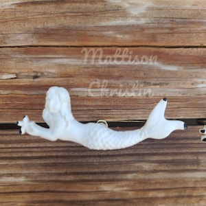 May include: White ceramic mermaid door knob with black eyes and a gold screw. The mermaid is swimming to the right with her tail curled up.