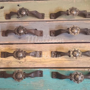 May include: A collection of rustic, decorative drawer pulls. Each pull features a dark brown, curved handle with a decorative silver-toned medallion. The medallion has a cross design surrounded by a floral pattern. The pulls are arranged on a weathered, multi-colored wooden surface.