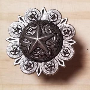 May include: A silver metal star-shaped decorative piece with a black center and a star design. The piece is surrounded by eight smaller stars.