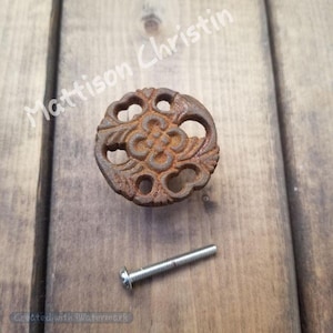 May include: A rusty brown, ornate cabinet knob with a floral design and openwork details. A silver screw is positioned next to the knob. The knob is on a wooden surface.