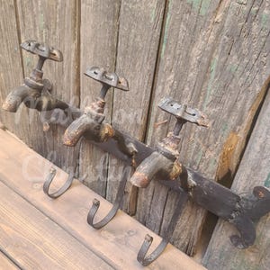 May include: Three rustic black metal hooks shaped like old-fashioned water faucets with a decorative scroll design at the end. The hooks are mounted on a wooden wall.