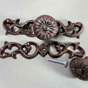 May include: Two ornate, bronze-colored drawer pulls with a decorative floral design. The pulls are attached to a matching, decorative, bronze-colored plate.