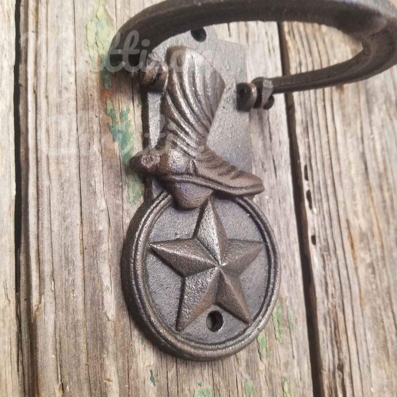Op de afbeelding: Een rustieke gietijzeren wandhaak met een cowboylaars en een ster. De haak is gemonteerd op een rechthoekige plaat en heeft een gebogen arm. De afwerking is donkerbruin en verweerd.