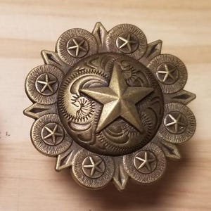 May include: A gold-toned metal star concho with a decorative design and eight surrounding stars. The concho is a popular embellishment for western wear and accessories.