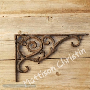 May include: A decorative, rusty brown metal shelf bracket with ornate scrollwork and a right-angle design. The bracket is mounted on a wooden surface. The text "Mattison Christin" is visible on the image.
