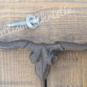 May include: A black cast iron bull skull door knob with horns. The knob is mounted on a wooden surface with a screw and washer.