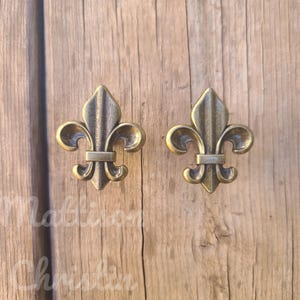 May include: Two antique brass fleur-de-lis cabinet knobs. The knobs are in a classic fleur-de-lis design with a detailed, aged finish. They are set against a wooden background, showcasing their decorative appeal. The knobs are likely for furniture or decorative use.