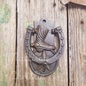 Op de afbeelding: Een gietijzeren deurklopper met een cowboylaars in een hoefijzer, met een ster aan de basis. De klopper is gemonteerd op verweerde houten planken, wat een rustiek, western-thema ontwerp laat zien.