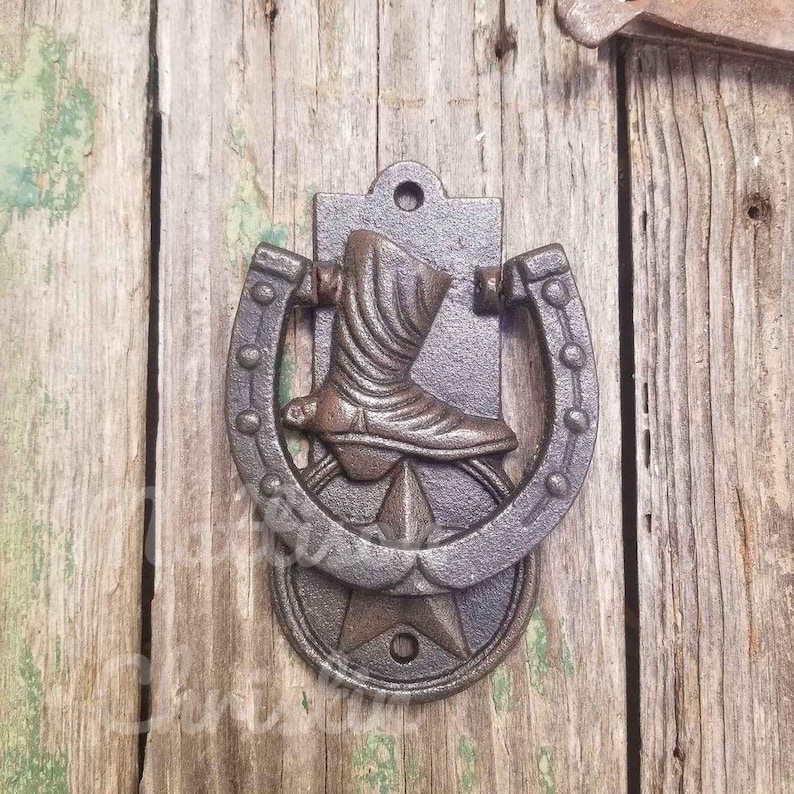 Op de afbeelding: Een gietijzeren deurklopper met een cowboylaars in een hoefijzer, met een ster aan de basis. De klopper is gemonteerd op verweerde houten planken, wat een rustiek, western-thema ontwerp laat zien.