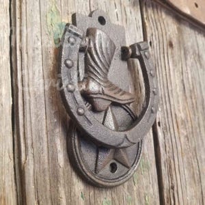 Op de afbeelding: Een rustieke deurklopper van gietijzer met een hoefijzerontwerp en een cowboylaars in het midden. De klopper is gemonteerd op een houten deur en heeft een stervormige basis. De kleur is een donker, verweerd bruin. Ongeveer 15 cm hoog.