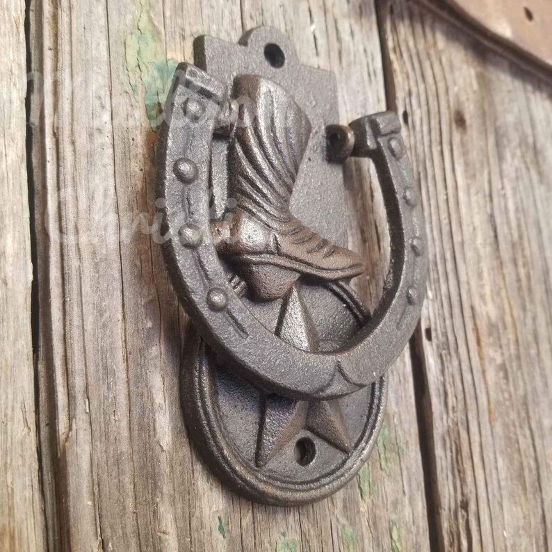 Op de afbeelding: Een rustieke deurklopper van gietijzer met een hoefijzerontwerp en een cowboylaars in het midden. De klopper is gemonteerd op een houten deur en heeft een stervormige basis. De kleur is een donker, verweerd bruin. Ongeveer 15 cm hoog.