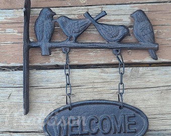 May include: Black cast iron welcome sign with four birds perched on a branch. The sign hangs from two chains and reads "WELCOME" in a decorative font.
