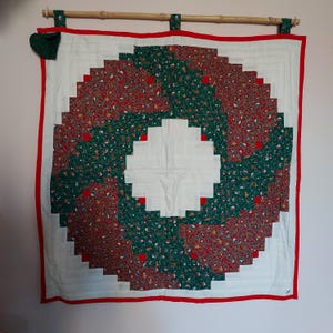 Peut inclure: Une tenture murale matelassée blanche avec un motif de couronne rouge et verte. La couronne est faite de carrés de tissu à motif floral.