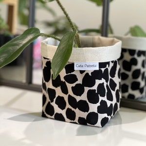 May include: A fabric pot with a black and white pattern, containing a green plant. The pot has a rolled-down top and a label that reads "Cutie Patootie". The pot is on a white surface. The pattern resembles a dalmatian's spots.