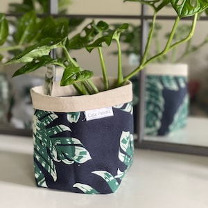 May include: A fabric plant pot with a navy blue background and a green and white tropical leaf pattern. The top edge is a natural beige colour. The pot contains a green plant with large leaves. The text "Cutie Patootie" is on a white label on the pot.