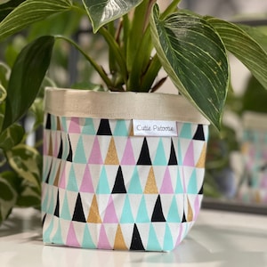 May include: A houseplant in a fabric pot with a geometric design of black, gold, pink, and turquoise triangles. The pot has a natural canvas top and a label that reads "Cutie Patootie."