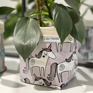 May include: A fabric plant pot featuring a repeating pattern of white unicorns with pink horns and manes on a pale purple backdrop. The pot contains a green leafy plant. The text "Cutie Patootie" is on a white label.