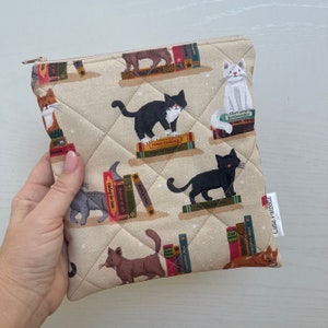 May include: Beige quilted zippered pouch with a repeating pattern of cats on books. The cats are black, white, grey, and brown. The books have titles such as "Home Cooking" and "Botanticals". The pouch is held in a hand.