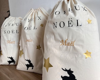 May include: Three off-white drawstring canvas sacks featuring the black text JOYEUX NOËL, gold star accents, and a black silhouette of a leaping reindeer. Each sack is personalised with a name in gold script: Matt, Louis, and Maël.
