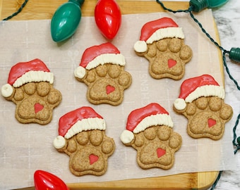 May include: Christmas-themed dog paw-shaped cookies, decorated with red Santa hats and heart-shaped accents. The cookies are arranged on parchment paper, with colorful Christmas lights in the background.