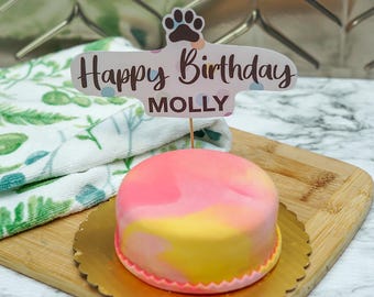 May include: A pink and yellow marbled cake with a Happy Birthday Molly topper. The cake is on a gold paper doily and a wooden cutting board.
