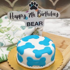 May include: A white dog cake with blue bone decorations and a Happy 7th Birthday Bear sign.