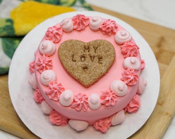 May include: A pink frosted cake with decorative pink frosting swirls and dollops. A heart-shaped cookie with the words "MY LOVE" is centered on top. The cake sits on a white plate.