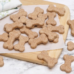 May include: A pile of brown dog biscuits in the shape of bones on a white parchment paper and a wooden cutting board.
