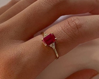 May include: A gold ring featuring a red emerald-cut gemstone and smaller clear gemstones on the band.
